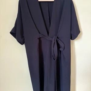 Svart omlottklänning från JW Make Way - Snygg svart omlottklänning från JW Make Way i storlek S. Klänningen har korta ärmar, knytband i midjan och en djup v-ringning. Stilren och enkel design som ger en cool och avslappnad vibe. 