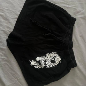 Svarta shorts med draktryck från SHEIN - Svarta mjukisshorts med vit drake tryckt på ena benet. De har elastisk midja med snörning för skön passform och är gjorda i mjukt bomullsmaterial. Perfekta för chill dagar eller träning. Snygg och edgy design som sticker ut. Fickor på sidorna