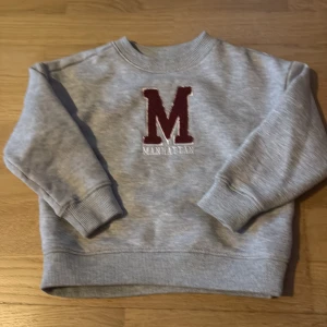 Grå sweatshirt med röd M från 157 - Grå sweatshirt från 157 med stor röd M och texten Manhattan broderat på bröstet. Tröjan har rund hals, ribbade muddar och är långärmad. Perfekt för dig som gillar college-stil och vill ha något bekvämt och snyggt.