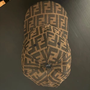 Fendi keps med FF-logga i brunt - Snygg keps från Fendi med det klassiska FF-monogrammet i brunt och svart. Kepsen har böjd skärm och justerbar rem baktill. Tillverkad i ett slitstarkt tyg med tydlig logotyp över hela ytan – perfekt för dig som gillar statement-plagg.