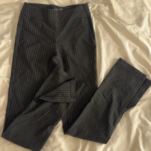 Svarta randiga kostymbyxor med slits - Snygga svarta kostymbyxor med tunna vita ränder. Byxorna är högmidjade och har bootcut-modell med slits nertill på benen och dragkedja på sidan av byxorna. Perfekta för en trendig och stilren look. 