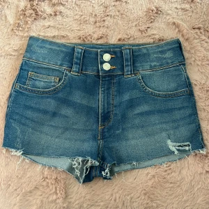 Blåa jeansshorts  - Snygga blå jeansshorts med mellanhög midja och råa, fransiga kanter. Shortsen har två knappar framtill, dragkedja och klassiska fickor både fram och bak. Perfekta för sommaren med sin slitna look och avslappnade vibe. Lappen är bortklippt men skulle tro att de är i storlek 158/164. Man kan även vika ner dem så att de blir low waist eftersom de är väldigt stretchiga, skriv om ni vill veta mått!