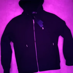 Svart hoodie från Polo Ralph Lauren - Svart hoodie från Polo Ralph Lauren med dragkedja och klassisk vit logga på bröstet. Tröjan har huva med snörning, långa ärmar och ribbade muddar. Perfekt för en chill och stilren look. Materialet är mjuk bomull som känns skönt mot huden.