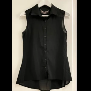 Rock Rebel svart ärmlös skjorta S - Svart ärmlös skjorta från Rock Rebel med klassisk krage och knappar framtill. Baksidan har en stor broderad döskalle i svart, vilket ger en edgy vibe. Skjortan är lätt och luftig, perfekt för dig som gillar en alternativ stil.