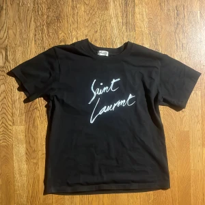 Svart Saint Laurent t-shirt - Säljer en snygg svart t-shirt ifrån Saint Laurent. Tröjan är helt ny. Fast det finns inga tags men den har inga defekter eller något annat! Priset är inte hugget i sten och kan gå ner vid snabb affär! Hör av dig vid minsta fråga!!
