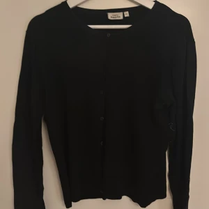 Svart kofta från KappAhl, storlek M - En klassisk svart kofta från KappAhl i storlek M. Koftan har rund halsringning, knäppning framtill med svarta knappar och långa ärmar. Perfekt att slänga över en t-shirt eller linne för en stilren look.