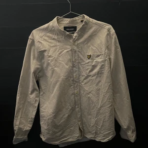 Beige skjorta (Lyle & Scott) - Stilren beige skjorta från Lyle & Scott i storlek L. Skjortan har klassisk krage, vita knappar, och en broderad logga på bröstet.