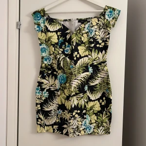 Blommig kortklänning med gröna toner - Snygg kortklänning med tropiskt blommönster i grönt, blått och beige mot svart bakgrund. Klänningen har en v-ringad hals och är ärmlös, perfekt för sommarens alla tillfällen. Materialet känns lätt och luftigt, och passformen är normal.