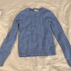 Blå kabelstickad tröja Holly & Whyte - Snygg ljusblå kabelstickad tröja från Holly & Whyte by Lindex. Tröjan har rund hals, ribbade muddar och ett klassiskt flätmönster över hela plagget. Perfekt för dig som gillar en stilren look. 