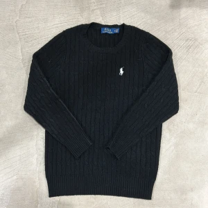 Svart stickad tröja Polo Ralph Lauren - Svart stickad tröja från Polo Ralph Lauren med klassisk kabelstickad design och broderad vit logga på bröstet. Rund halsringning och långa ärmar. Perfekt för dig som gillar stilrena och tidlösa plagg med premiumkänsla.
