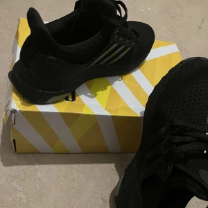 Svarta Adidas Ultraboost sneakers - Säljer ett par svarta Adidas Ultraboost sneakers i storlek 38. Skorna har en stickad ovandel och klassiska tre ränder på sidan. Snygg och sportig design med platt sula och snörning. Perfekta för dig som gillar stilrena och bekväma sneakers.