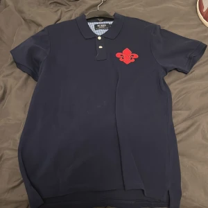 Mörkblå Morris pikétröja L - Snygg mörkblå pikétröja från Morris med röd logga framtill och stora röda bokstäver på ryggen. Klassisk krage, tre knappar och korta ärmar. Materialet är mjuk bomull som känns skönt mot huden. Perfekt för dig som gillar stilren design med en sportig touch. väldigt bra skick