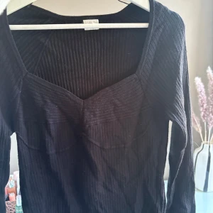 Svart ribbad topp  - Svart långärmad topp från Stories med ribbad struktur och markerad byst. Snygg, figurnära passform och djupare urringning. 