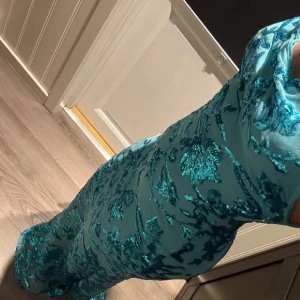 Turkos glittrig långklänning med blommor - Säljer en turkos långklänning med smala axelband och v-ringning. Klänningen har ett glittrigt blommönster i paljetter över hela tyget och sitter figurnära. Perfekt för dig som vill sticka ut med en färgstark look!