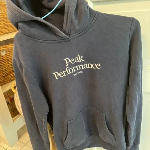 Mörkblå hoodie från Peak Performance - Snygg mörkblå hoodie från Peak Performance med vit broderad logga på bröstet. Klassisk känguruficka framtill och huva. Perfekt för chill dagar eller när du vill ha en sportig look.