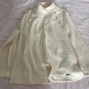 Vit cape-kappa/poncho - Stilren vit cape-kappa med hög krage och dubbla rader av guldiga knappar framtill. Jackan har slitsade fickor. Strl XS/S