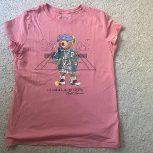 Rosa Polo Bear t-shirt från Ralph Lauren - Snygg rosa t-shirt från Ralph Lauren med ikoniska Polo Bear-trycket på bröstet. T-shirten har rund halsringning och korta ärmar. Tillverkad i mjuk bomull, köpt ifrån små hjärtan i Höllviken för 700 kr.