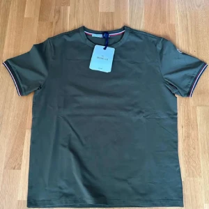 Grön Moncler t-shirt med detaljer - Snygg grön t-shirt från Moncler med korta ärmar och diskreta rödblå-vita ränder på ärmsluten. Klassisk rund halsringning och Moncler-logga på ärmen. Perfekt för en clean och stilren look.