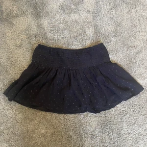 Svart prickig kortkjol Gina Tricot - Supersöt svart kortkjol från Gina Tricot med små glittriga prickar och veckad midja. Kjolen har en lätt volang och är gjord i ett tunt, flowy material som känns skönt mot huden. Perfekt för dig som gillar en lekfull och trendig stil.