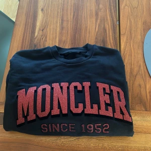 Svart Moncler sweatshirt - Svart sweatshirt från Moncler med stor röd  text 'MONCLER SINCE 1952' på bröstet. Klassisk rund halsringning och långa ärmar. Tillverkad i mjuk bomull för en skön och avslappnad vibe. Aldrig använd. 