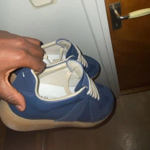 Blå sneakers från Maison Margiela - Säljer ett par stilrena sneakers från Maison Margiela i marinblått och beige. Skorna har en klassisk rund tå, beige snörning och en platt gummisula. Materialet är en mix av mocka och skinn, vilket ger en lyxig känsla. Perfekt för dig som gillar minimalistisk design och snygga detaljer.