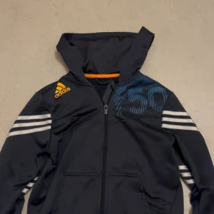 Mörkblå Adidas kofta med huva - Mörkblå  från Adidas med huva och hel dragkedja. har vita ränder på ärmarna, gult Adidas-tryck på axeln och blått grafiskt tryck på bröstet. Tillverkad i polyester, perfekt för träning eller chill. Snygg och sportig look med normal passform.