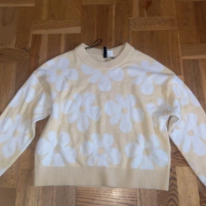 Beige blommig stickad tröja Divided - Superfin beige stickad tröja från Divided med stora vita blommor över hela framsidan och ärmarna. Tröjan har rund halsringning, ribbade muddar och en avslappnad passform. Perfekt för dig som gillar en söt och trendig look!