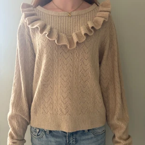 Beige stickad tröja med volang - Supersöt beige stickad tröja med volangdetalj runt halsen. Tröjan har ett diskret hålmönster och långa ärmar med ribbade muddar. Perfekt för dig som gillar en romantisk och trendig look.