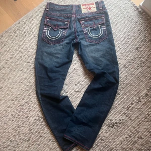True Religion jeans med rosa sömmar - Säljer ett par blå jeans från True Religion med ikoniska bakfickor och kontrasterande rosa och vita sömmar. Klassisk femficksmodell med raka ben och detaljerade nitar. Har dock inga knappar på bak fickorna.☺️