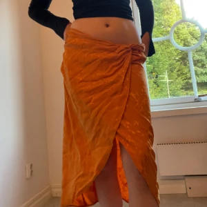 Orange omlottkjol från Gina Tricot - Snygg orange omlottkjol från Gina Tricot i storlek 34. Kjolen har en knytning framtill, subtilt blommönster och en slits. Materialet är glansigt och lent. 