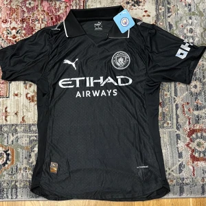 Manchester City svart Puma Third kit matchtröja - Svart Manchester City fotbollströja Spelarversion från Puma med klassisk krage och vita detaljer. Tröjan har korta ärmar, broderad klubbmärke och Puma-logga på bröstet samt Etihad Airways-tryck. Tillverkad i lätt funktionsmaterial (Ultraweave) för maximal komfort.