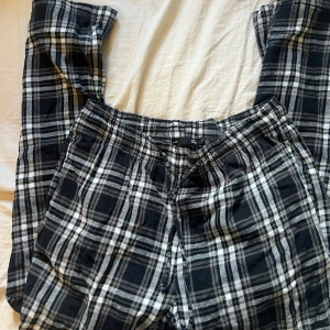 Svarta rutiga byxor från 57 - Säljer ett par svarta och vita rutiga byxor från Lager  157 med resår i midjan. De är tillverkade i mjukt bomullstyg och har en avslappnad passform, perfekta för chill dagar hemma eller när du vill vara bekväm men ändå se snygg ut.