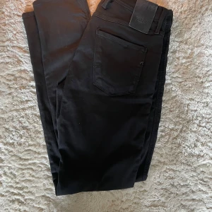 Svarta Replay Anbass jeans slim fit - Svarta jeans från Replay, modell Anbass med slim fit. Klassisk femficksdesign, svarta sömmar och diskret Replay-logga på fickan. Snygg och stilren look som passar till det mesta. Mjukt och stretchigt material för extra komfort.