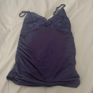 Mörklila spetsdetaljerad linnetopp - Supersöt marinblå linnetopp med smala axelband och v-ringad spets upptill. Toppen har en liten rosett framtill och rynkade sidor för extra snygg passform. Perfekt för dig som gillar romantiska detaljer och mjuka material.