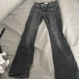 Grå bootcut jeans från Gina Tricot - Säljer ett par grå bootcut jeans från Gina Tricot med snygga slitningar och markerade sömmar. Jeansen har två framfickor med lock och knapp, samt klassiska bakfickor. Perfekta för dig som gillar en retro vibe och vill ha något som sticker ut.