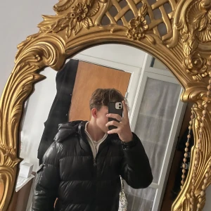 Svart Moncler pufferjacka med huva - Sjukt grisch pufferjacka från Moncler med glansig yta och huva. Jackan har dragkedja framtill, två sidofickor med dragkedja och Moncler-logga på ärmen. Perfekt för kalla dagar och har en riktigt schysst streetstil.