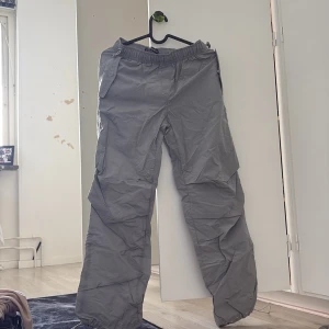 Grå cargopants från New Yorker XXs - Snygga grå cargopants från Sister XX med elastisk midja och dragsko vid benslut. Byxorna har flera fickor och är tillverkade i ett lätt syntetmaterial som ger en avslappnad och loose passform. Perfekta för en chill och street-inspirerad look.