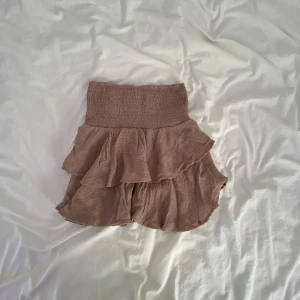 Beige volangkjol från SHEIN - Supersöt beige kortkjol från SHEIN med bred smock i midjan och dubbla volanger. Kjolen är luftig och har en flowy känsla, perfekt för varma dagar. Materialet känns lätt och skönt mot huden. Enkel att matcha med crop tops eller stickade tröjor.