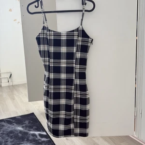 Svartvit rutig kort klänning - Snygg svartvit rutig klänning med smala axelband och figurnära passform. Klänningen är kort och har en klassisk fyrkantig urringning. Perfekt för dig som gillar en stilren och trendig look med lite edge.