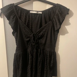 Svart blus med snörning och volang - Supersöt svart blus från lager 157 med volangärmar och snygg snörning framtill. Toppen har en lätt rynkad struktur och v-ringning, perfekt att styla med jeans eller kjol. Luftig och skön modell med feminina detaljer.