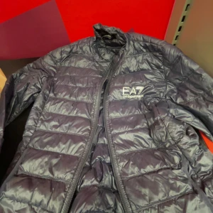 Svart pufferjacka från EA7 Emporio Armani - Säljer en svart pufferjacka från EA7 Emporio Armani med snyggt quiltad design och logga på bröstet. Jackan har dragkedja framtill och står ut med sin sportiga look. Två st väldigt små hål vid högra armen. Jag är 182 och de passar mig helt perfekt. Priset kan diskuteras🤝
