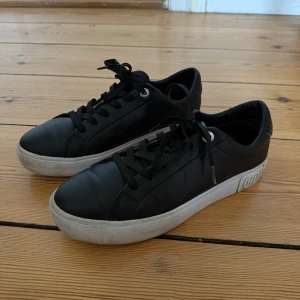Svarta sneakers från Tommy Hilfiger - Snygga svarta sneakers från Tommy Hilfiger med vit platt sula och klassisk snörning. Skorna har rund tå och är tillverkade i skinn med metallögla och logga på hälen. Perfekta för dig som gillar stilrena och tidlösa sneakers.