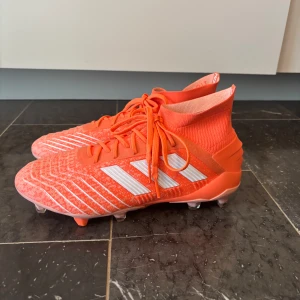 Adidas Predator Coral fotbollsskor - Säljer ett par Adidas Predator fotbollsskor i en riktigt snygg orange färg med vita detaljer och mönster. Skorna har snörning, strumpliknande skaft och en platt sula med dobbar för gräsplan. Perfekt för dig som vill sticka ut på planen och ha grym kontroll på bollen.