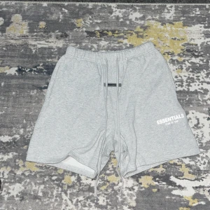 Grå Essentials shorts med snörning - Snygga grå shorts från Essentials Fear of God med elastisk midja och långa snören framtill. Mjuk bomullskänsla, relaxed fit och diskret logga på benet. Perfekta för chill dagar och streetwear-stil. Bakficka för extra funktion.