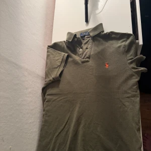  pikétröja från Polo Ralph Lauren - Säljer en klassisk olivgrön pikétröja från Polo Ralph Lauren med orange broderad logga på bröstet. Tröjan har krage, knappar vid halsen och korta ärmar. Perfekt för en stilren och avslappnad look.