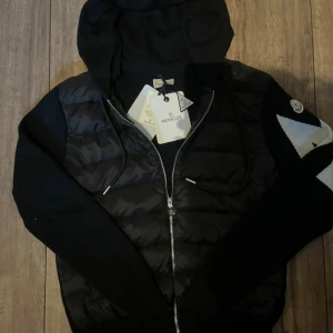 Svart cardigan från Moncler M - Svart pufferjacka från Moncler med huva och dragkedja framtill. Jackan har quiltad front i blank polyester och stickade ärmar med Moncler-logga på vänster arm. Snygg, sportig look med snörning vid huvan och ribbade muddar. Perfekt för dig som gillar streetstyle.