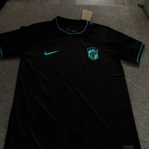 Svart Brasilien Nike fotbollströja - Snygg svart Brasilien fotbollströja från Nike med Dri-Fit material. Tröjan har korta ärmar och coola turkosa detaljer på krage och ärmslut. Brasilien-emblemet och Nike-loggan i turkos ger en modern vibe. Perfekt för dig som vill sticka ut på planen eller gatan.