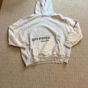 Grå hoodie Tommy Hilfiger - Snygg grå hoodie från Tommy Hilfiger med stor svart logga och texten 'New York' på framsidan. Tröjan har huva, magficka och ribbade muddar vid ärmar och nederkant. Perfekt för chill dagar och streetstyle. Mjuk bomullskänsla och relaxed fit.