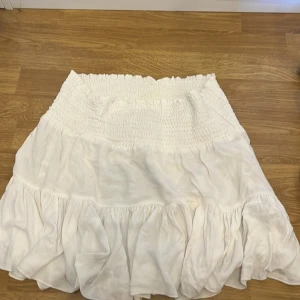 Vit volangkjol från lager 157, storlek L - Superfin vit kortkjol från lager 157 med bred smock i midjan och volangig nederdel. Kjolen är luftig och har en lätt struktur i tyget som ger en chill vibe. Perfekt för varma dagar när du vill ha något bekvämt och snyggt.