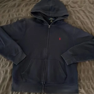 Mörkblå hoodie från Polo Ralph Lauren - Mörkblå hoodie från Polo Ralph Lauren med dragkedja och klassisk röd logga broderad på bröstet. Tröjan har huva, känguruficka och långa ärmar med muddar. Perfekt för dig som gillar stilren och enkel streetwear. om du hr några frågor så var inte rädd att skriva till mig